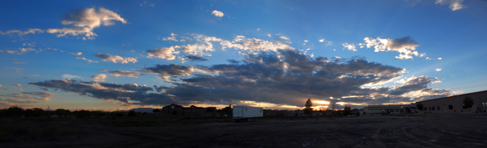 [Several photos stitched together showing a large cloud mass through whith the setting sun shines through openings in the clouds. Blue sky is seen all around the cloud mass. Top of the clouds are white while the underside is dark grey.]